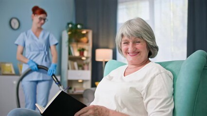 portrait of a happy old woman with a book smiling and looking at the camera background of female social worker with a vacuum cleaner in her hands