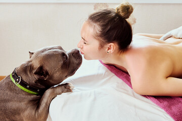 An American Bully dog kisses young woman who is lying on massage table in a massage parlor.