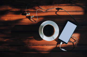 Black smartphone with white screen, dark headphone and cup of coffee lies on a brown wooden...