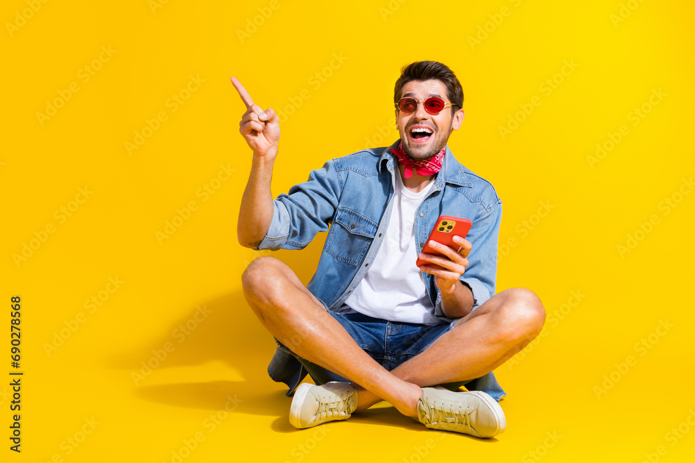Poster full body photo of excited man dressed denim shirt sit with smartphone look directing at empty space