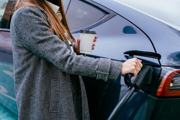 Unrecognizable woman holding cup of coffee charging electro car at the electric gas station....