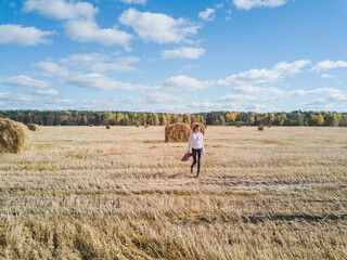 Woman enjoys walking and spending weekend on harvested farm field under blue sky. Tourist explores field with straw bales against forest