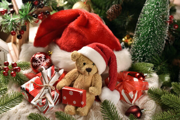 Teddy bear toy in Santa hat sitting with presents in front of Christmas tree and big red hat