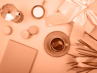 There is a coffee pot on the table, a cup of coffee, a notebook and flowers. The background is peach color.