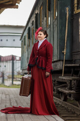 Fototapeta premium A beautiful girl in a burgundy suit of the last century and a hat with a veil stands with a suitcase in her hands near an old steam locomotive.Vintage portrait of the last century, retro journey