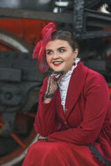 A beautiful girl in a burgundy suit of the last century and a hat with a veil is sitting on a suitcase near an old steam locomotive.Vintage portrait of the last century, retro journey