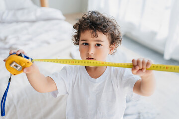 Curly confused handsome little boy holding measuring roulette, wants growing faster. Children psychology. Pretty kid puzzled by his small height. Spanish kid with tape measuring at home showing height - obrazy, fototapety, plakaty