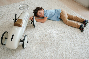 Upset little caucasian boy laying on carpet at home with toy car, angry, tired, with empty space....