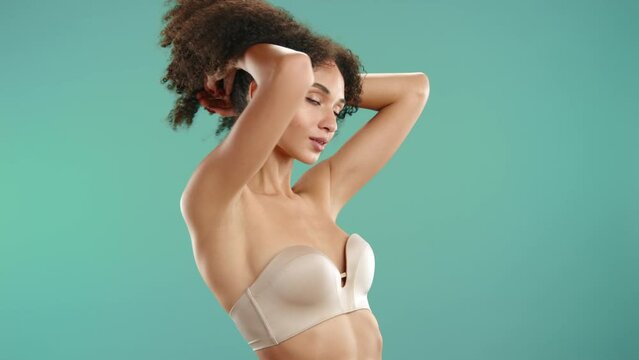 Attractive young model with beautiful body wearing ultimate strapless nude bra closing eyes, raising dark fluffy curls posing on blue green background. High quality 4k footage