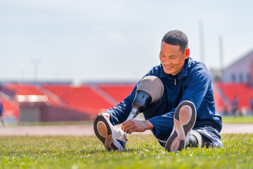 Sport man athlete with a prosthesis on his leg sit on grass field to tie shoelaces near the track...
