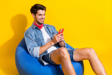 Portrait of clever man dressed denim shirt red scarf sit on bean bag look at smartphone isolated on...