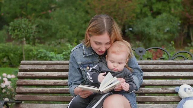 Happy mother reading book to while relaxing on bench. Smiling woman spending leisure time with cute toddler. She is wearing denim jacket at park during summer.