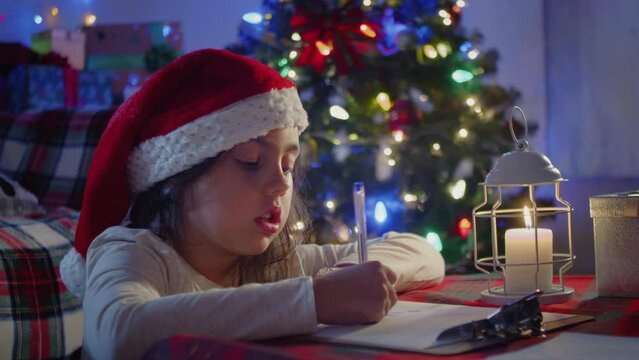 Funny girl in a Santa hat writes a letter to Santa near the Christmas tree