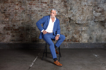 A handsome middle-aged man in a blue formal suit poses sitting in a chair. Portrait of a businessman. A handsome middle-aged man poses on a brick background with a place to copy