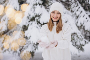 A happy woman in knitted mittens poses against the background of a snowy pine tree in winter