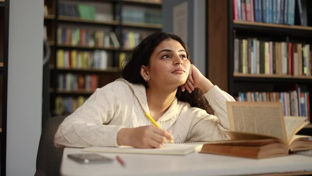 Smiling young pretty woman student dreaming searching inspiration looking for solution idea answer sitting at desk in the university library Beautiful curly girl thinking about great weekend indoors