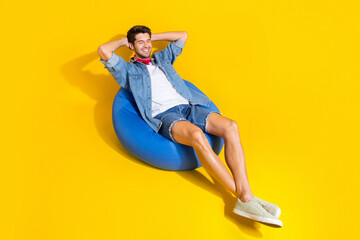 Portrait of good mood man dressed denim shirt sit on bean bag at vacation arms behind head isolated on vivid yellow color background