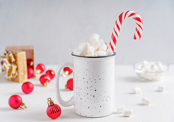 Homemade sweet cocoa drink or hot chocolate with marshmallow topping decorated with striped candy cane served in cup or mug on white wooden table with bright red christmas baubles and golden gift box