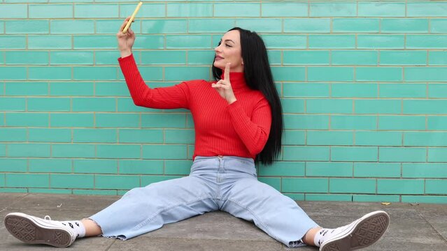 young adult woman wearing a red sweater resting sitting on the floor while taking a selfie photo with her cell phone