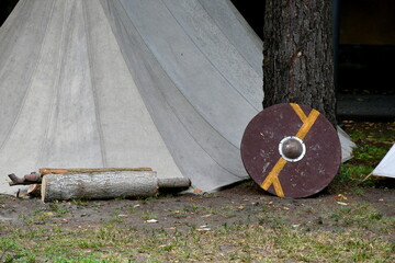 A close up on a wooden targe or shield located next to a big tree and a white tent made out of cloth spotted during a medieval fair or festival organized in Poland in summer