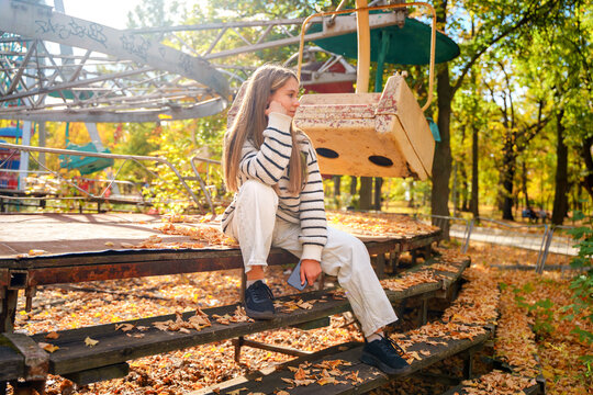 A teenage girl in an abandoned amusement park in autumn, a stylish girl sits near the carousel. Togliatti, Russia - 30 Sep 2023