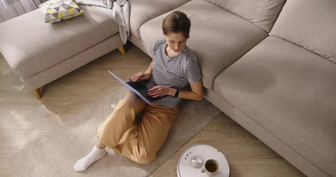 Attractive calm freelance female working from home taking mug cup from robot vacuum cleaner. Home sweet home. Smart technologies. People spending time usefully instead of households. Cinematic AD