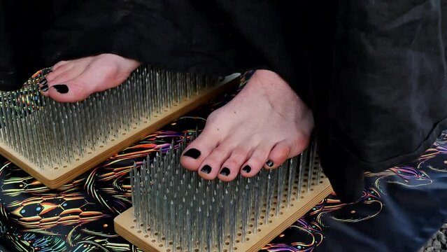 Close-up of a woman's bare feet with black nails stepping on the sharp nails of a Sadhu. A barefoot woman with a black pedicure and a black dress stood on the Sadhu board where there are tall nails