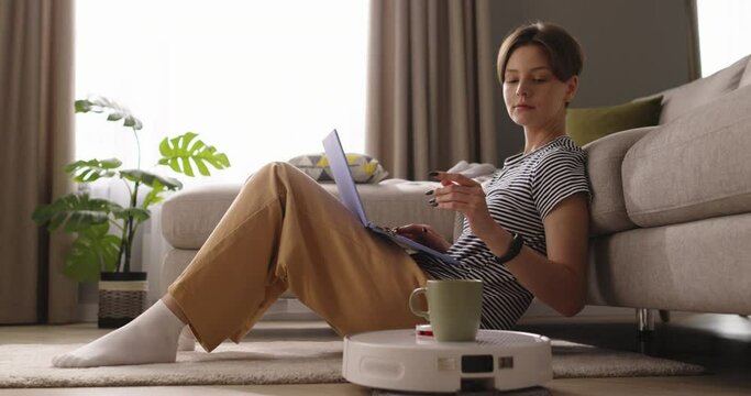Young attractive caucasian woman working on laptop. Robot vacuum cleaner delivering coffee. Concept of hygiene, household gadgets and robots at modern life. Smart technologies. Cinematic advertising