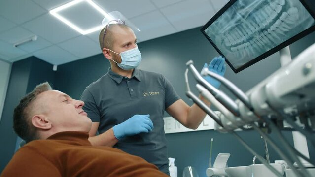 Professional dentist in medical mask, gloves in fully equipped dental office consulting middle-aged patient using display with X-ray of jaw and teeth. High quality 4k footage