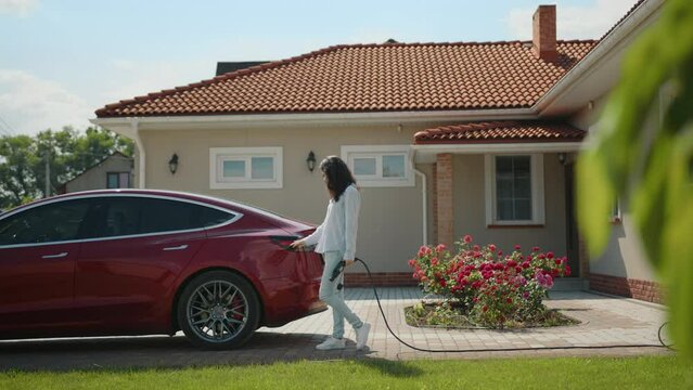 Young cute brunette woman puts red electric car on charge in the yard at home. Electric car stands near modern one-storey house with well-kept territory. High quality 4k footage