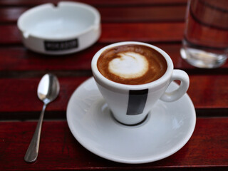 americano cup on saucer with spoon, ashtray, glass of water on red wooden coffee shop table (espresso, milk, macchiato) european style (cappuccino)