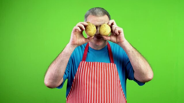 Cheerful senior chef covering eyes with potatoes.