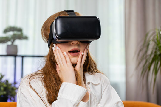 Teenager red hair girl use virtual reality futuristic technology VR app headset helmet to play simulation 3D 360 video game, watching film movie at modern home apartment. Child in goggles sits on sofa