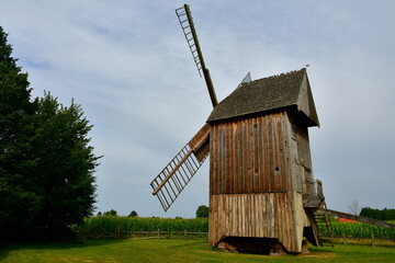 A view of a big mill with wooden blades spotted on a lush field next to some crops and a vast forest or moor seen on a cloudy summer day on a Polish countryside during a hike