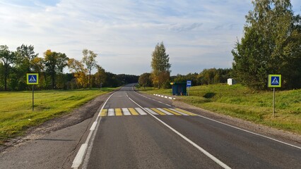 Fototapeta na wymiar There is a crosswalk marked with markings and signs near the bus stop on an asphalt road with shoulders among grassy meadows and mixed forest. In the fall, yellow foliage appears on the trees. Sunny