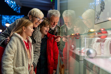 Happy grandparents with grandchildren looking at store window. An elderly man and woman are taking their grandchildren Christmas shopping. - obrazy, fototapety, plakaty
