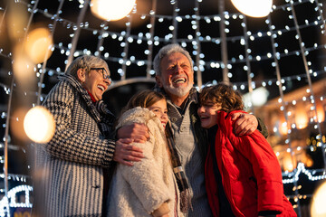 Overjoyed senior couple having fun with granddaughter and grandson surrounded with Christmas lights...