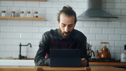 Bearded young man sitting in kitchen, using app with tablet device, surfing on internet