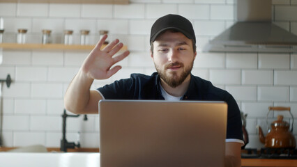 Young adult man in a hat sitting in the kitchen, participating in a remote video chat by waving, video chatting with friends or family with a laptop