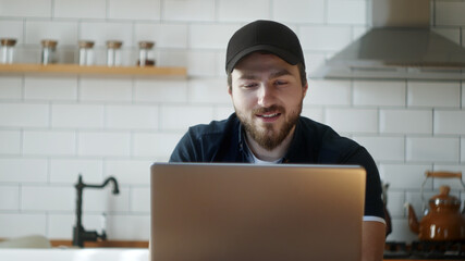 Handsome young man in a hat sitting in the kitchen, participating in a remote video chat, video chatting with friends or family with a laptop