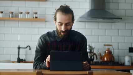 Bearded young adult man sitting in kitchen, using app with tablet device, surfing on internet,...