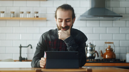 Bearded young adult man sitting in kitchen, watching video or show with tablet device, surfing on...