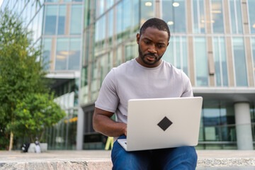 African American man freelancer using laptop typing chatting on urban street in city. Guy having virtual meeting online chat conference. Entrepreneur programmer working on laptop outdoors. Remote work