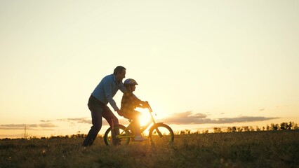 Persistent boy in helmet learns to ride bike under careful guidance of considerate daddy. Loving father holds bicycle to ensure safety of little son. Family weekends on grassy meadow at sunset - obrazy, fototapety, plakaty