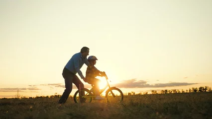 Deurstickers Persistent boy in helmet learns to ride bike under careful guidance of considerate daddy. Loving father holds bicycle to ensure safety of little son. Family weekends on grassy meadow at sunset © DREAM INSPIRATION
