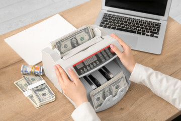 Woman putting dollar banknotes into cash counting machine at table in office, closeup - obrazy, fototapety, plakaty