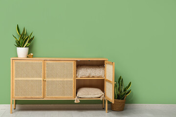 Modern light wooden chest of drawers with houseplants and pillows near green wall in room