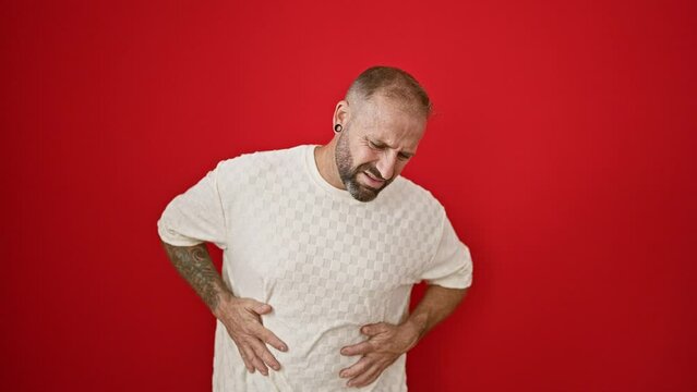 Worried young man, cool yet serious, suffering stomach ache, desperately standing over an isolated red background, exuding unhappiness and confusion.