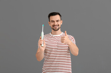 Handsome man with electric toothbrush showing thumb-up on grey background