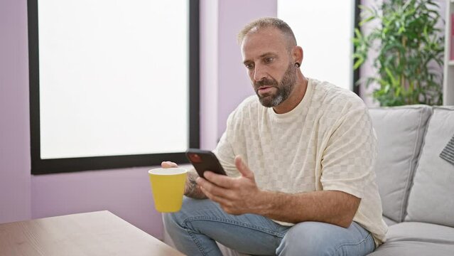 Surprised young man, sitting relaxed on sofa at home, drinking morning espresso from a mug, looking at his smartphone screen with amazed expression